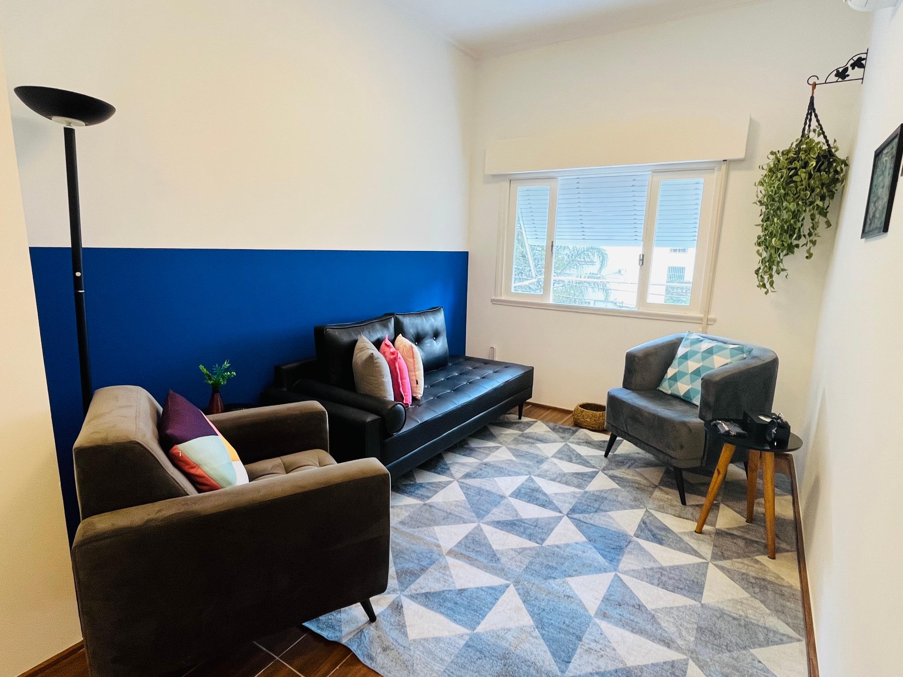 Sala 4 com parede azul, divã de couro preto de almofadas coloridas, duas poltronas e tapete geométrico
