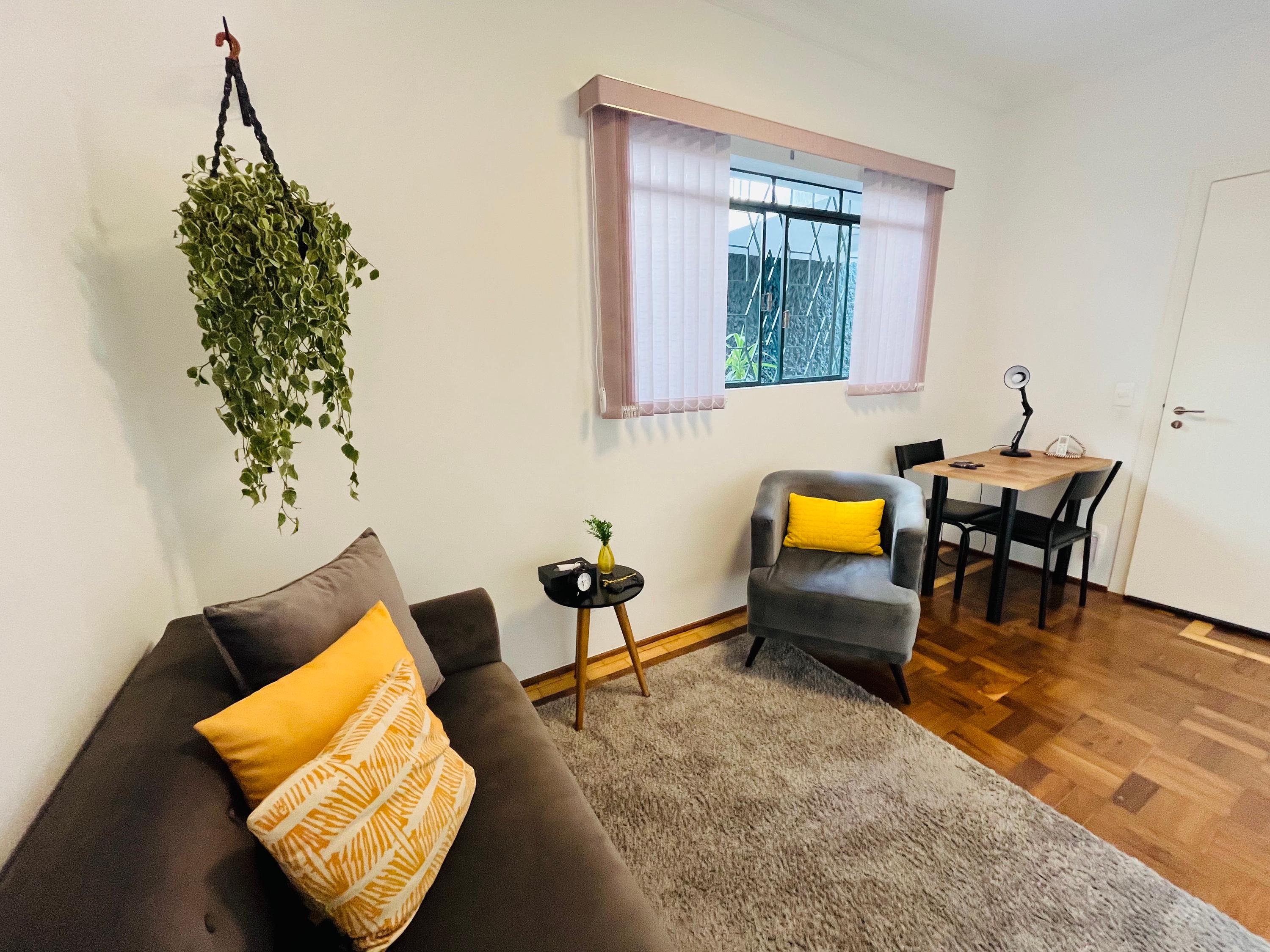 Sala 1 vista do sofá, com poltrona cinza, planta pendurada e escrivaninha junto à porta