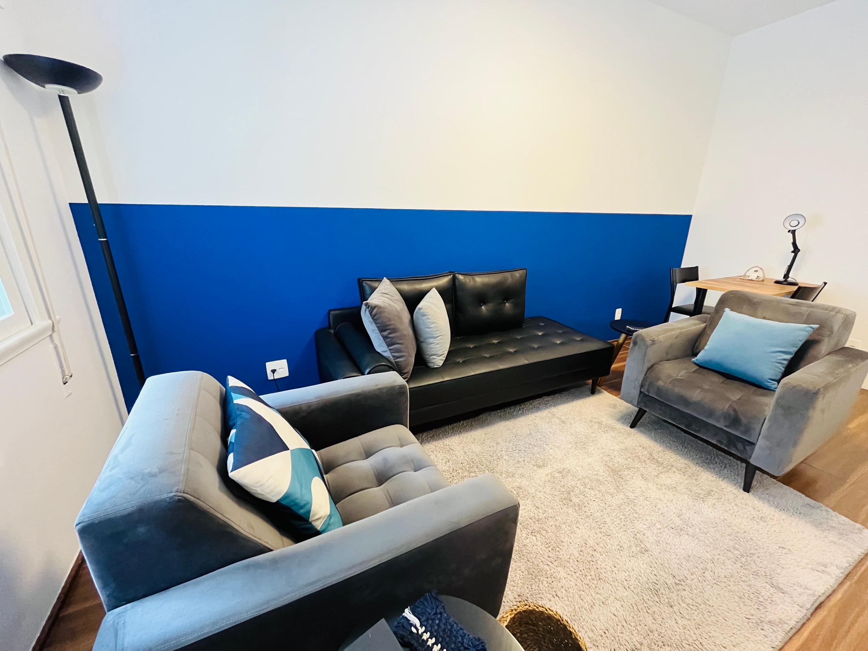 Sala 5 com parede azul, divã de couro preto, duas poltronas cinza e escrivaninha ao fundo