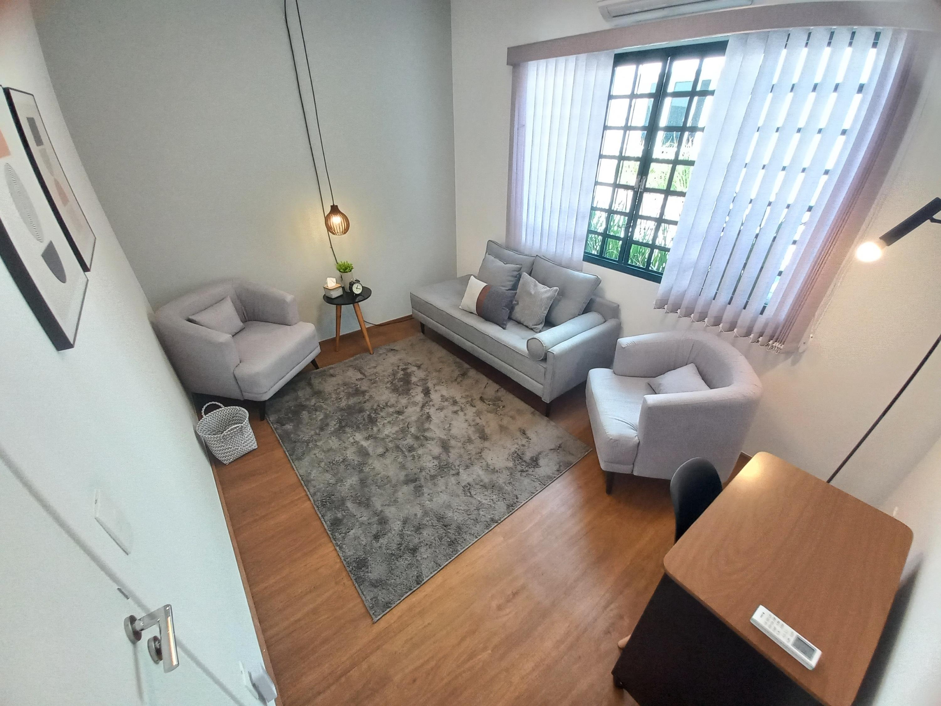 Sala 7 com divã cinza entre duas poltronas, tapete e janela com persiana vertical